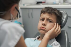 A young boy sits in the dentist's chair with his hand on his cheek, looking distressed as he has tooth pain